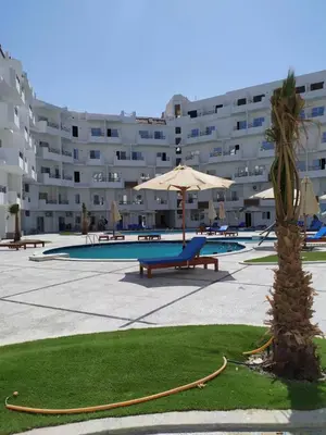a pool with an umbrella and a palm tree in front of a building at Tiba View flat near pool in Hurghada
