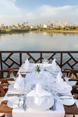 a table with white plates and napkins and a view of a river at Farouz El Nil I Nile Cruise - Every Monday from Luxor for 07 & 05 Nights in Ţawd