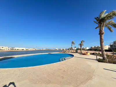a swimming pool next to a beach with a palm tree at Stylish 1BR Ground With Free Pool & Lagoon Access In Gouna in Hurghada
