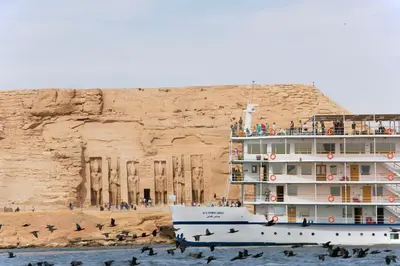 a cruise ship in the water next to a cliff at Mövenpick Prince Abbas Lake Nasser Cruise, Every Monday Four Nights From Aswan, Every Friday Three nights From Abu-Simbel - Including Excursions and Sightseeing in Aswan